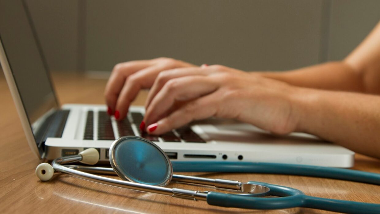 Hands typing on a laptop on a wooden desk, with a stethoscope lying beside it