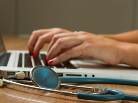 Hands typing on a laptop on a wooden desk, with a stethoscope lying beside it