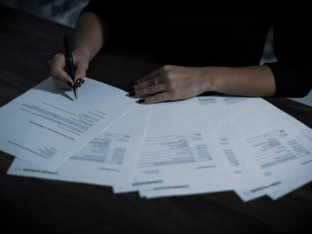 A person holding a pen over several financial statements spread across a dark table